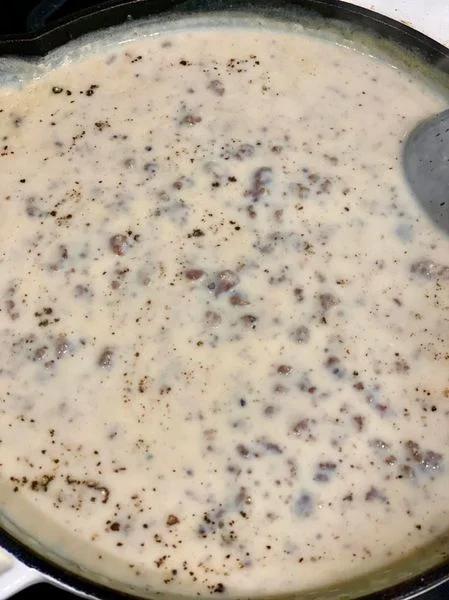 Milk gravy with sausage cooking in a skillet, topped with black pepper.