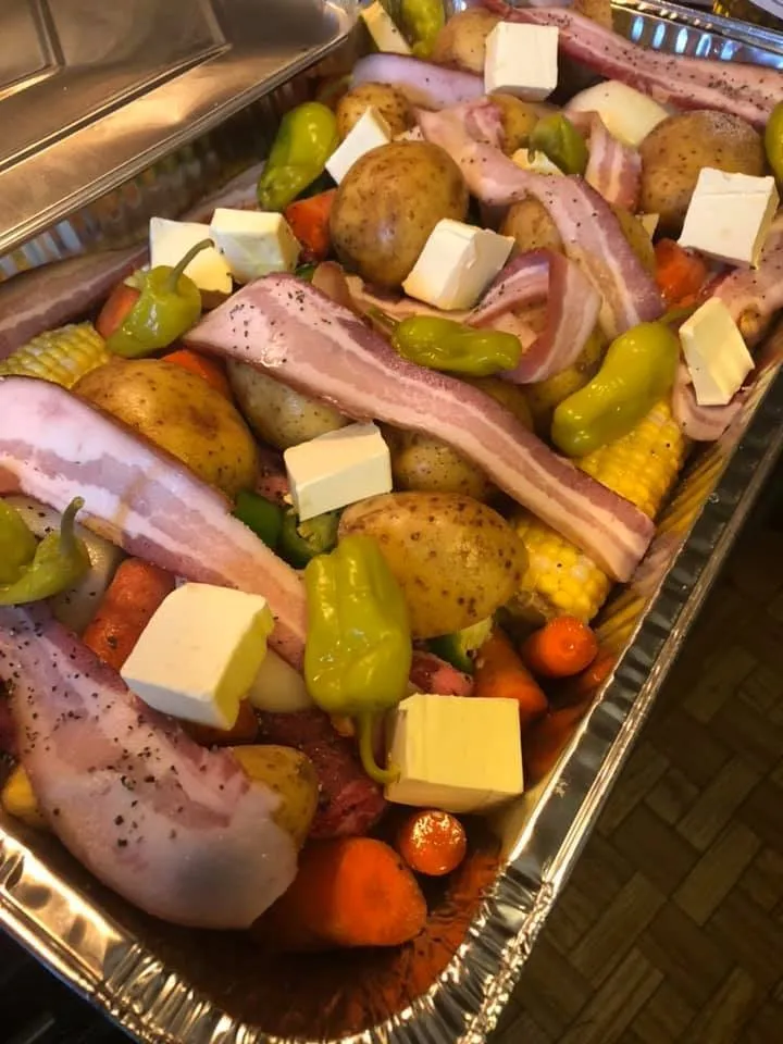 Uncooked easy chuck roast dinner layered in a foil pan with potatoes, carrots, corn, bacon strips, butter cubes, and pepperoncini peppers ready for the oven.