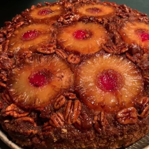 A freshly baked Upside-Down Candied Pecan Pineapple Cake served on a round tray, displaying a glossy topping of caramelized pineapple rings, cherries, and glazed pecans.