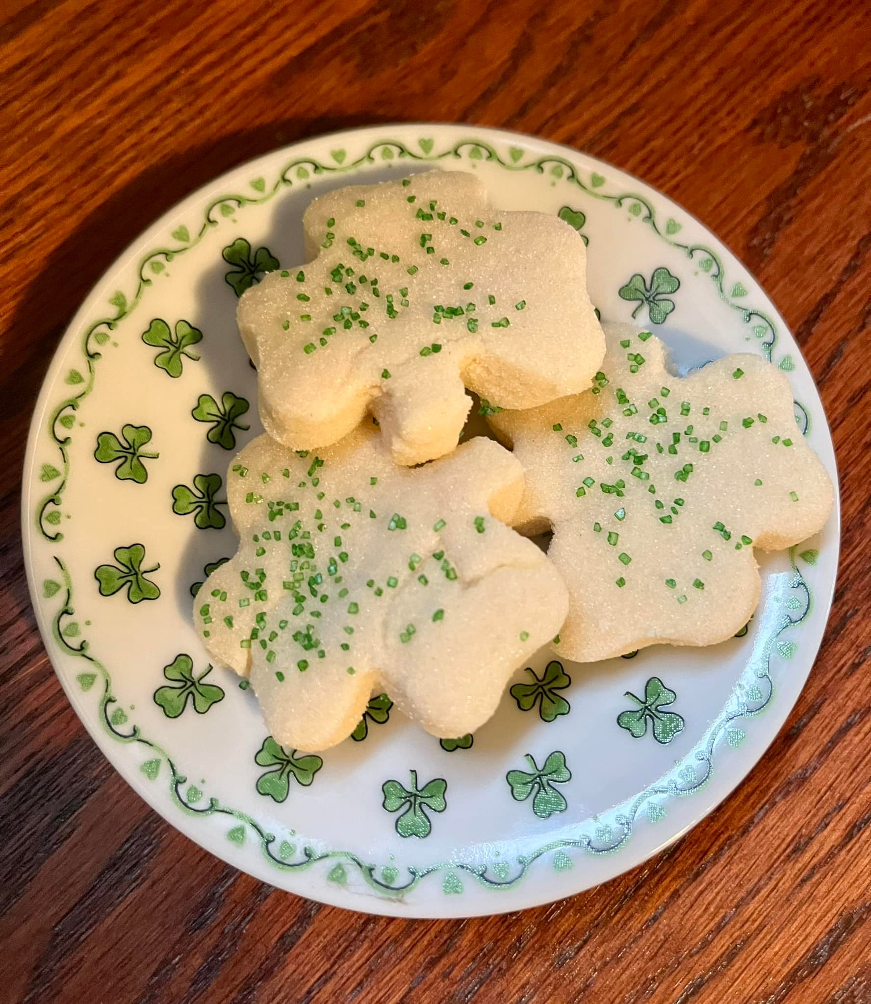 Three shamrock-shaped shortbread cookies on a clover-decorated plate, topped with green sugar.