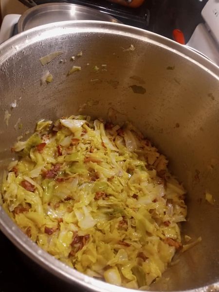 Chopped cabbage frying in a stainless steel pot with crispy bacon and butter.