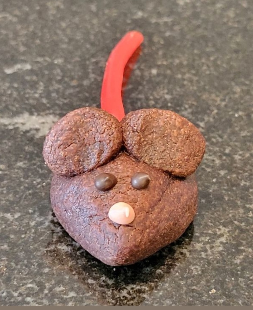 A baked chocolate mouse cookie with licorice tail, chocolate eyes, and a pink candy nose, sitting on a dark countertop