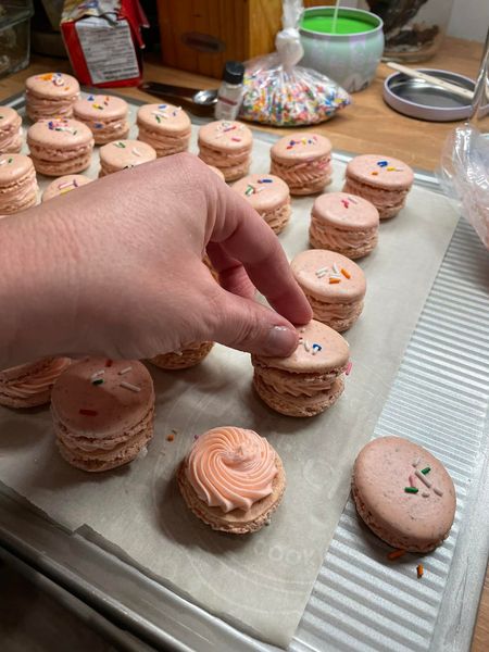 A hand placing the top shell on a macaron filled with pink buttercream frosting and garnished with rainbow sprinkles.