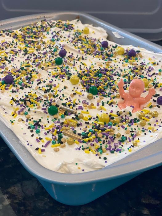 A King Cake Poke Cake decorated with purple, green, and gold sprinkles and topped with a festive baby figurine