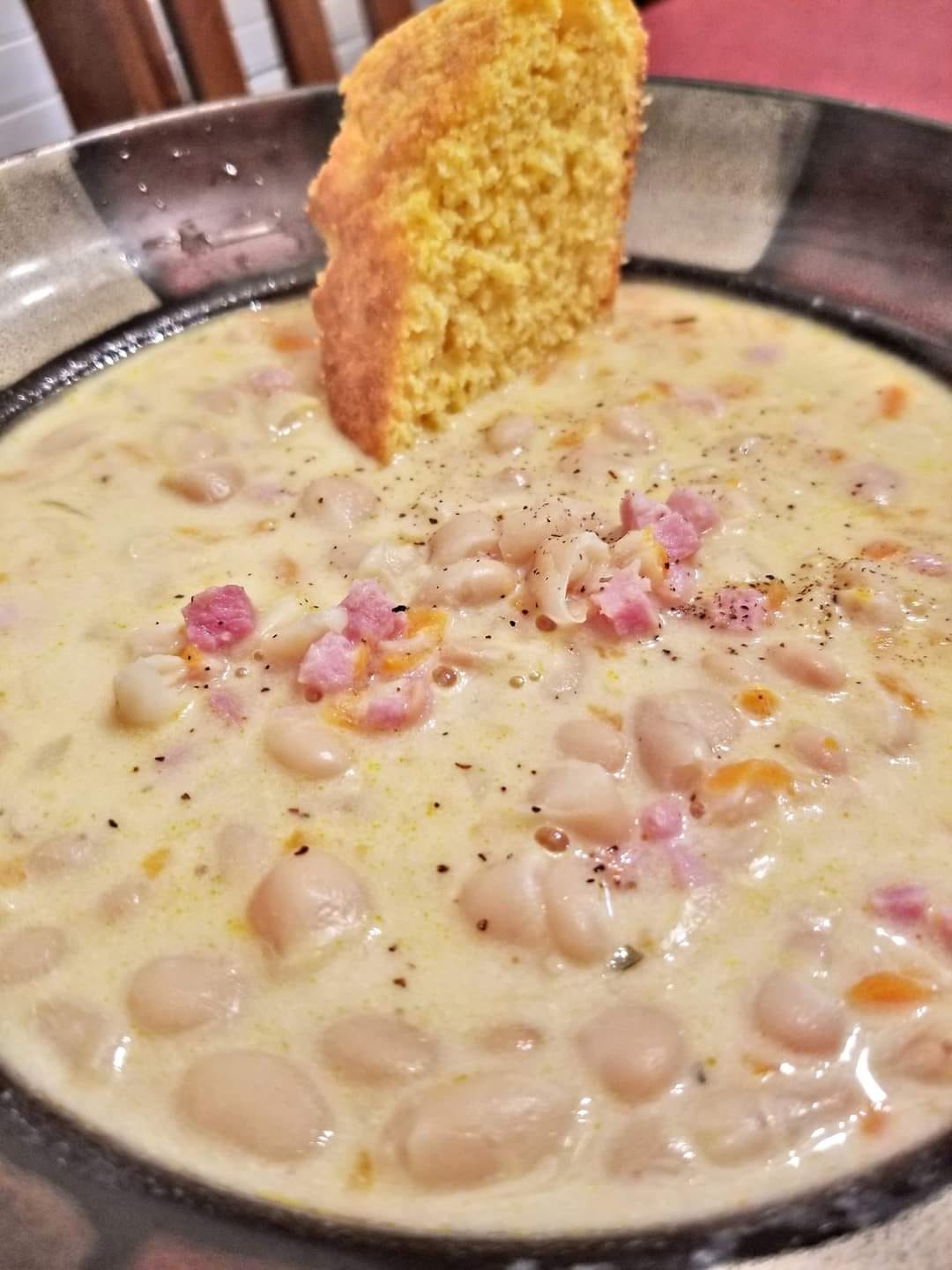 A bowl of creamy white bean chowder with diced ham and a slice of cornbread for dipping.