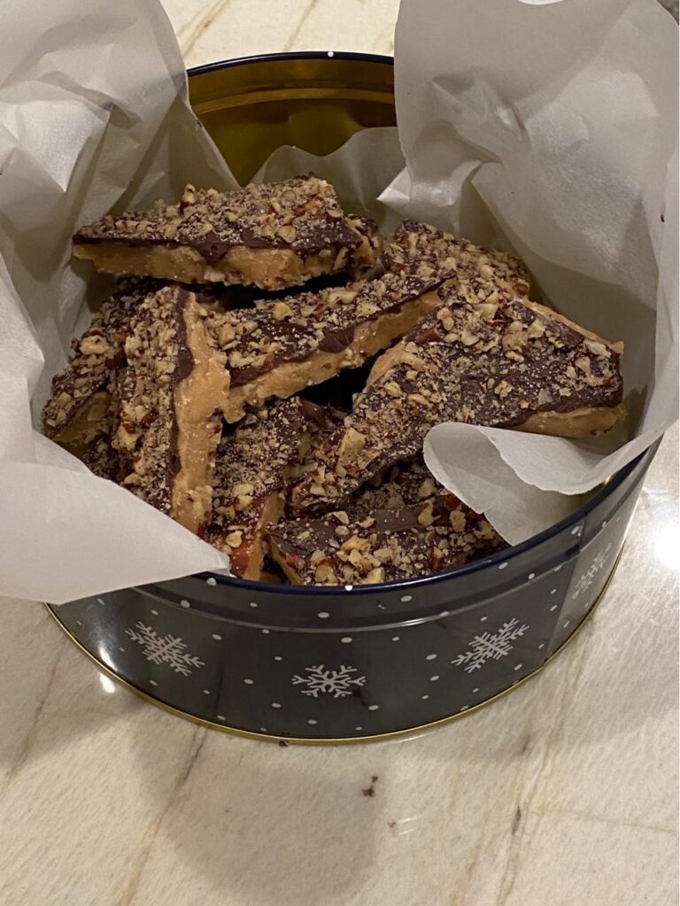 Homemade English toffee in a festive tin lined with parchment paper, topped with chopped nuts and chocolate.