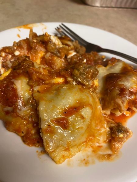 A serving of cheesy, meaty baked ravioli on a white plate, with a fork in the background. The dish is covered in tomato sauce and gooey melted cheese