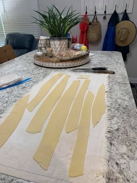 Rolled-out egg noodle dough cut into wide strips on a floured cloth, ready to be sliced into noodles