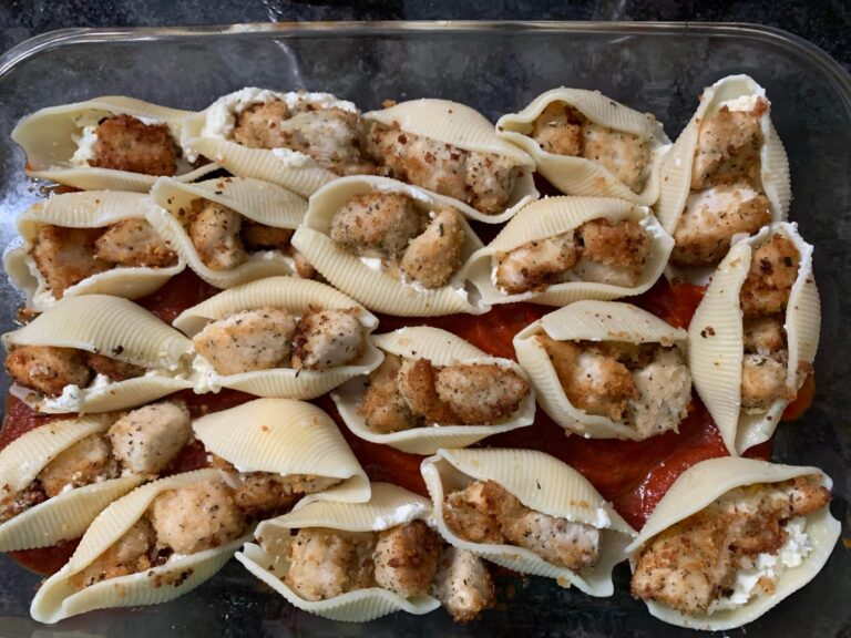 A glass baking dish filled with large pasta shells stuffed with ricotta and crispy breaded chicken, topped with marinara sauce and shredded mozzarella before baking.