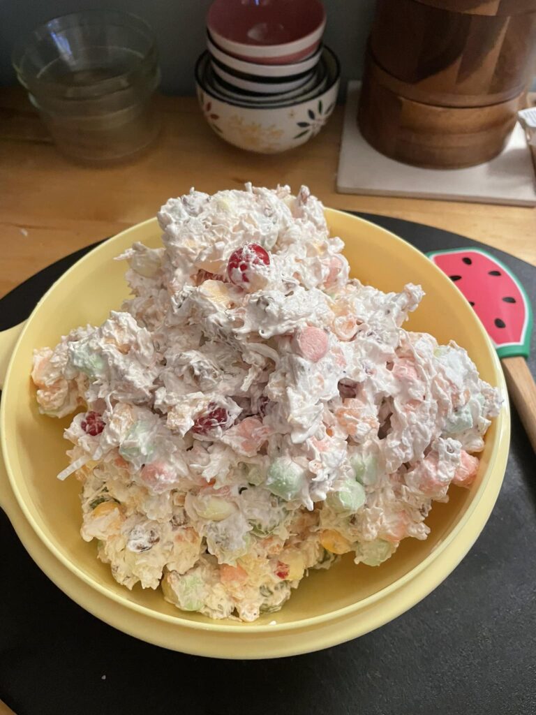 A large bowl of creamy Ambrosia Salad, filled with marshmallows, cherries, grapes, and coconut.