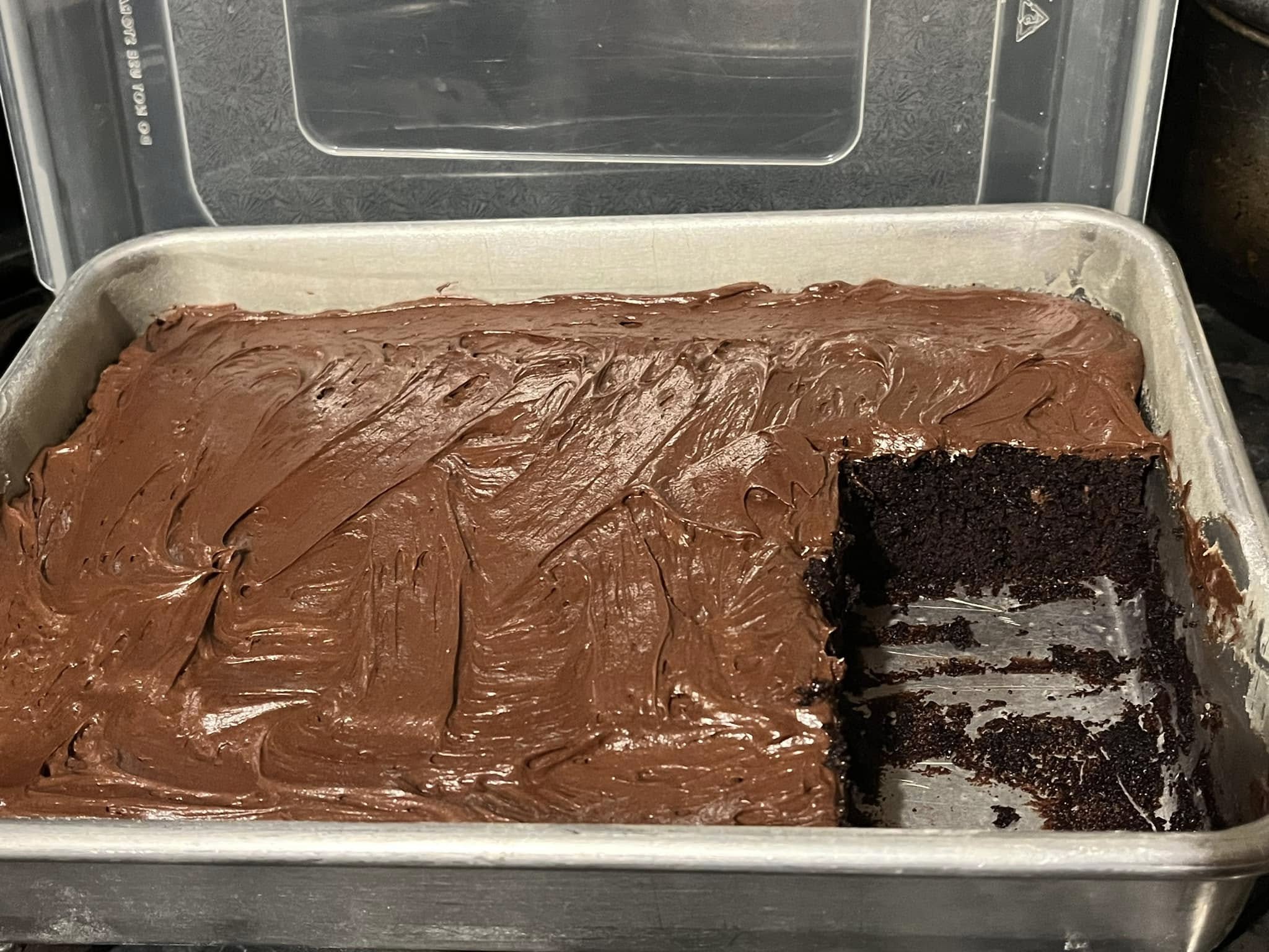 A freshly baked chocolate cake in a rectangular pan, covered with rich chocolate frosting, with one piece cut out to reveal its moist texture.