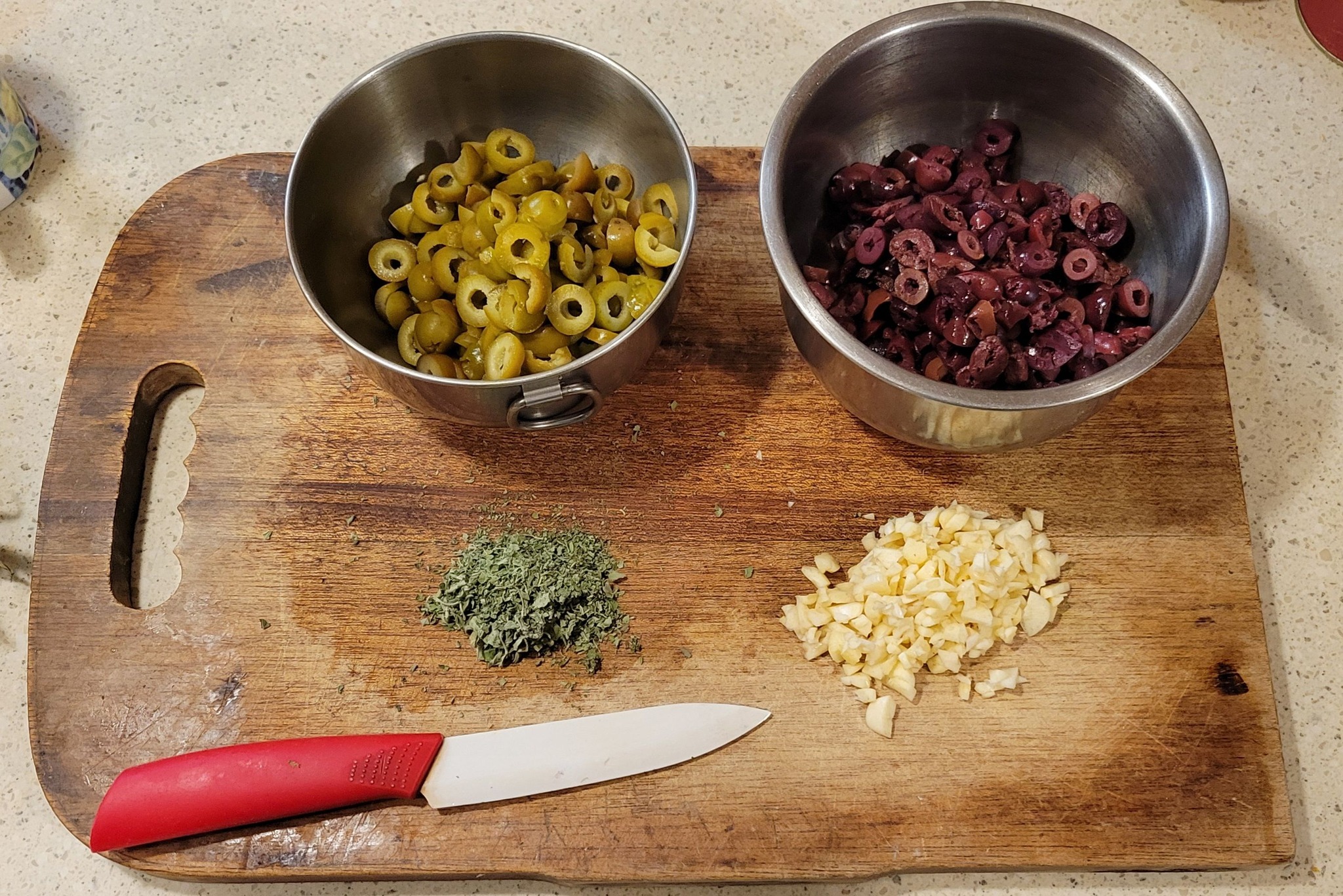 A wooden cutting board with chopped green and Kalamata olives, minced garlic, dried oregano, and a red-handled knife.