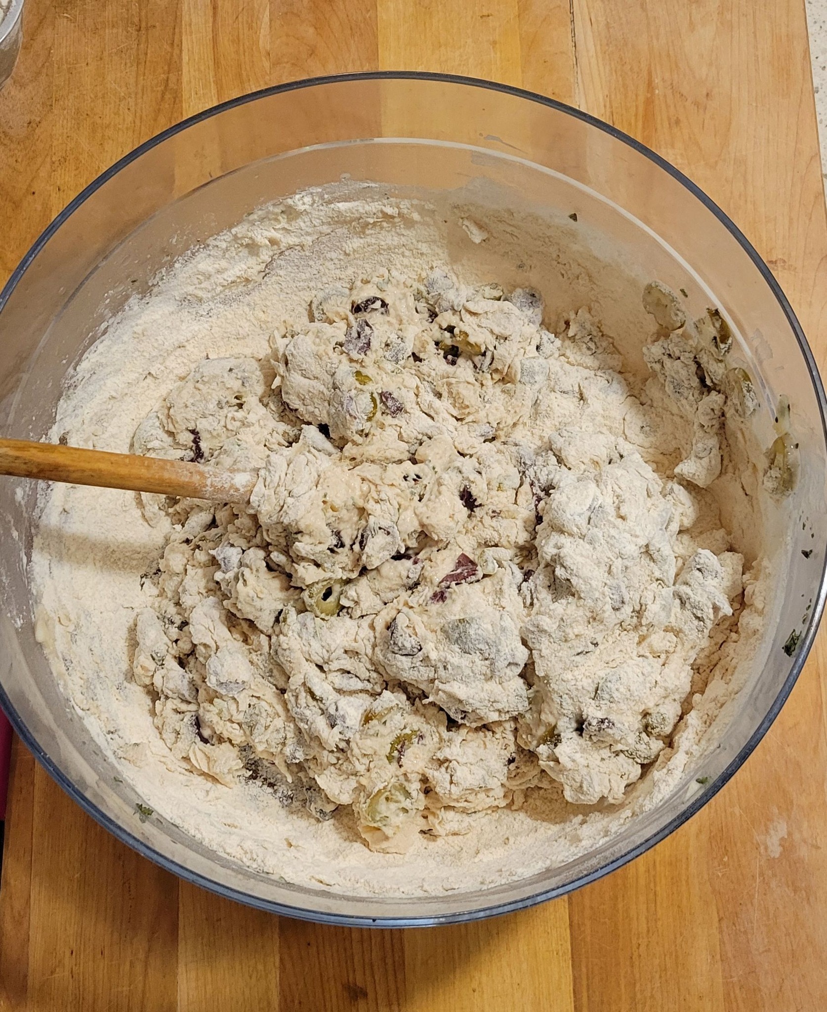 A thick, slightly sticky dough with olives and flour in a mixing bowl being stirred with a wooden spoon.