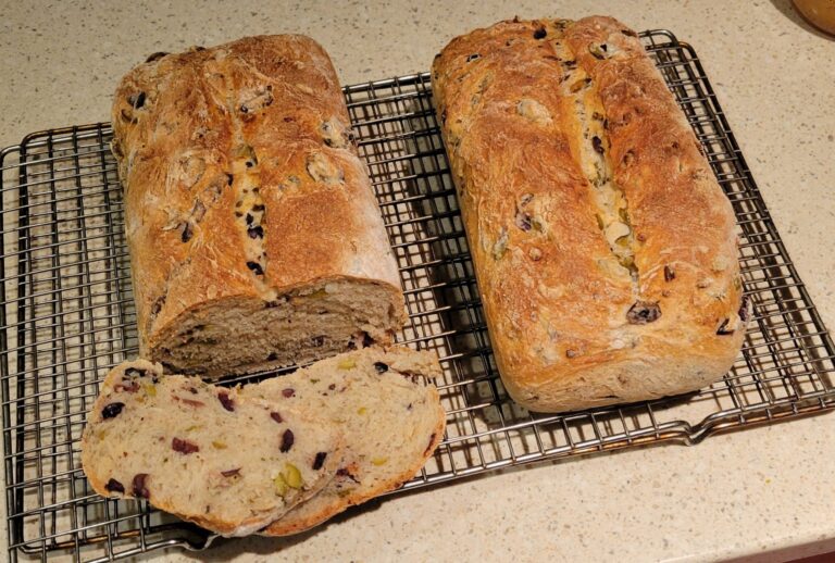 Two golden-brown loaves of olive loaf on a cooling rack, one partially sliced open to reveal a soft interior with olives.