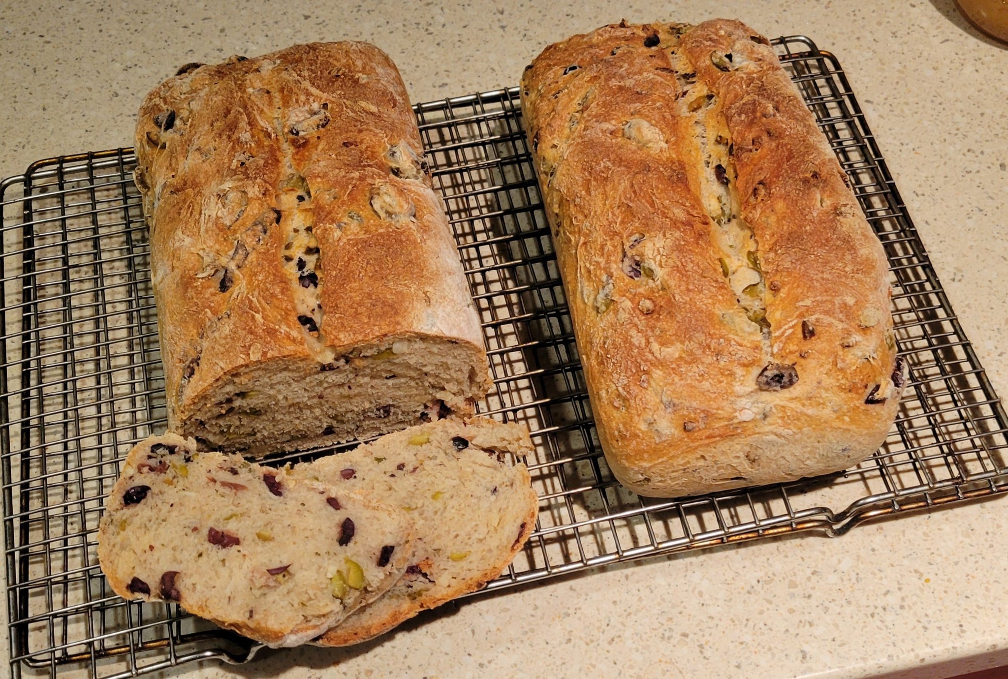 Two golden-brown loaves of olive loaf on a cooling rack, one partially sliced open to reveal a soft interior with olives.