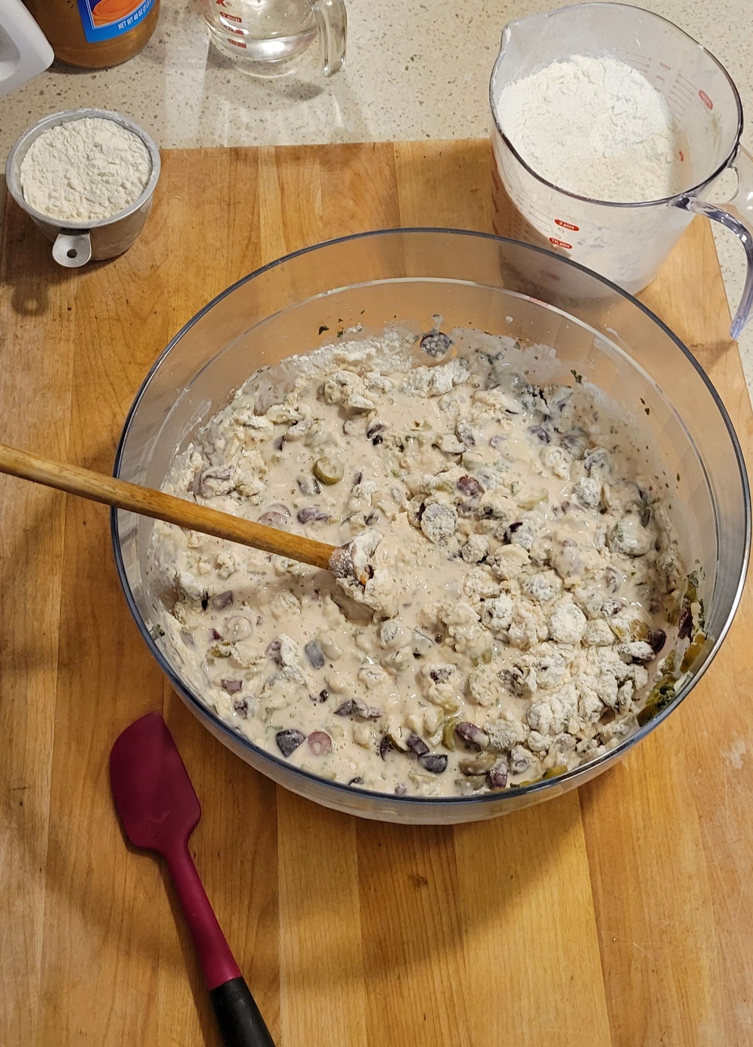 A large mixing bowl with flour being gradually added to a wet dough mixture containing olives, garlic, and oregano.