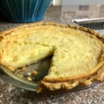 Buttermilk pie in glass pie dish with one slice missing, showcasing creamy texture.