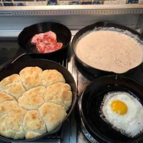 Southern breakfast with cast iron skillet biscuits, creamy sausage gravy, ham slices, and a fried egg.