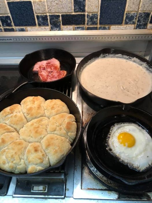 Southern breakfast with cast iron skillet biscuits, creamy sausage gravy, ham slices, and a fried egg.