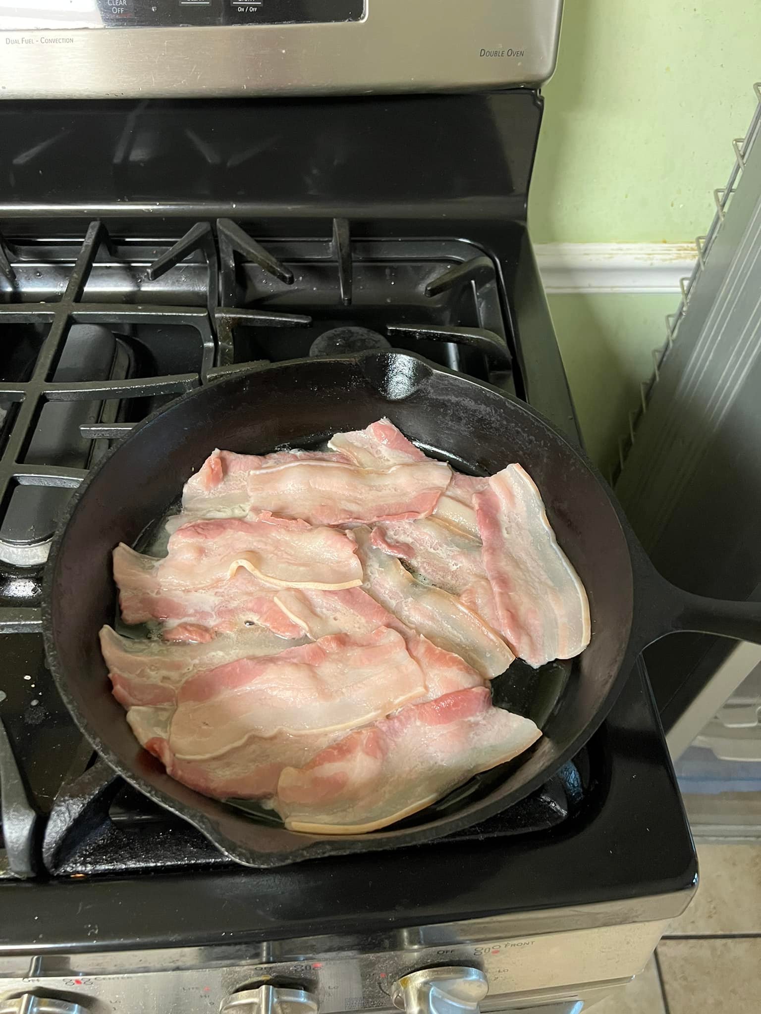 Raw bacon sizzling in a black cast iron skillet on a stovetop.