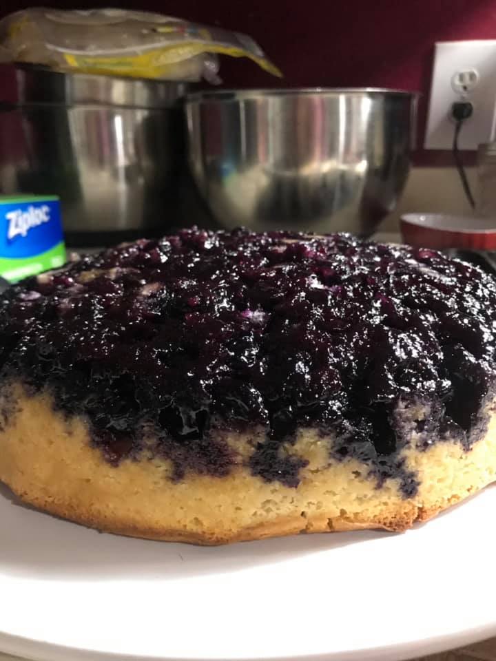 Whole blueberry upside-down cake flipped onto a white plate, topped with glistening berries.