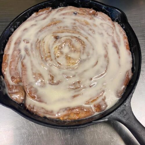 A cast iron skillet with a giant cinnamon roll swirled tightly and topped with creamy coffee-maple icing.