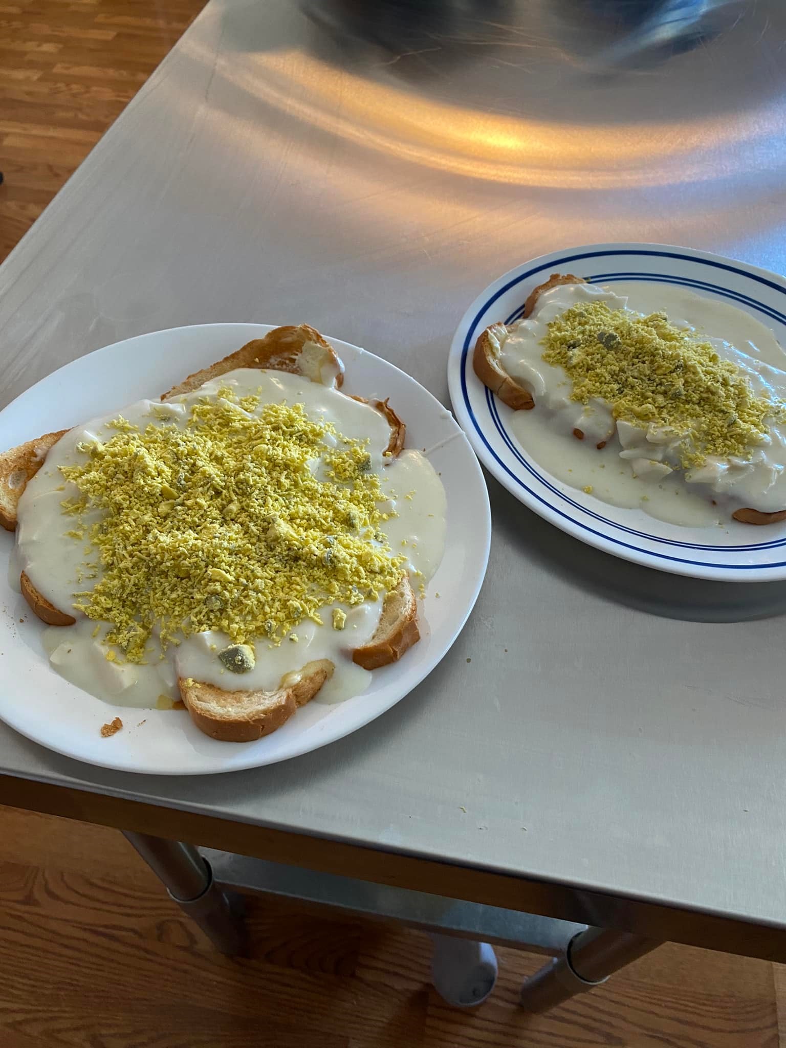 [Two plates of eggs à la goldenrod served on white toast, topped with grated yolk, and paired with orange juice on a metal table
