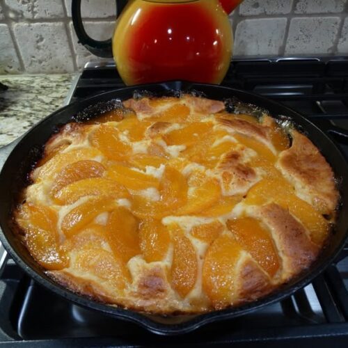 Golden brown peach skillet cobbler baked in a cast iron pan, topped with sliced canned peaches and served warm straight from the oven.