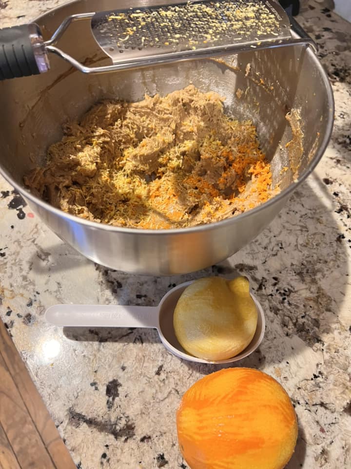 Mixing lemon zest, orange zest, and grated apple into homemade fruitcake batter in a stainless bowl.