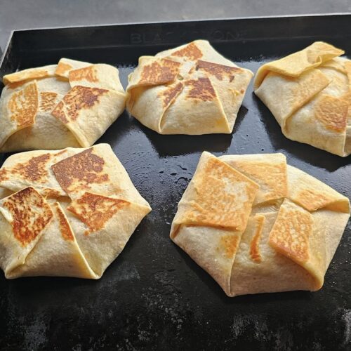 Five crunchwrap supremes crisping on outdoor griddle.