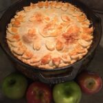 Golden brown cast iron skillet apple pie cooling on counter with fresh apples
