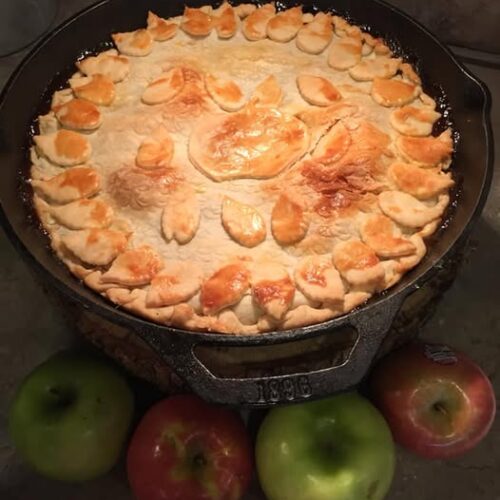 Golden brown cast iron skillet apple pie cooling on counter with fresh apples