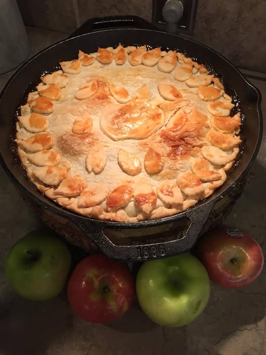 Golden brown cast iron skillet apple pie cooling on counter with fresh apples