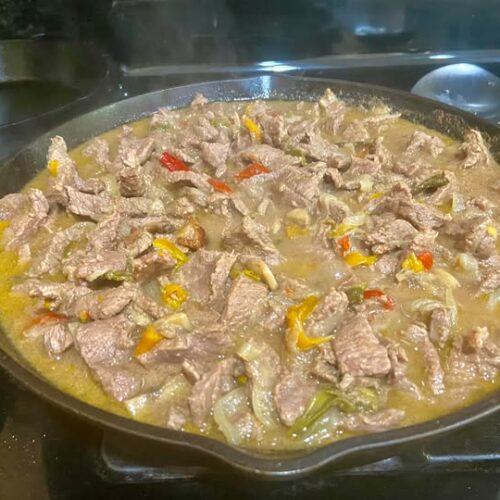 cast iron skillet filled with beef strips, peppers, and onions for homemade Philly cheesesteak
