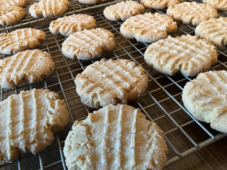 dozens of soft peanut butter cookies cooling on racks with sugar sparkle, surrounded by peanut butter and chocolate ingredients