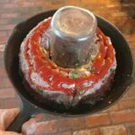 Raw meatloaf shaped around a jar in cast iron skillet, topped with Sriracha ketchup