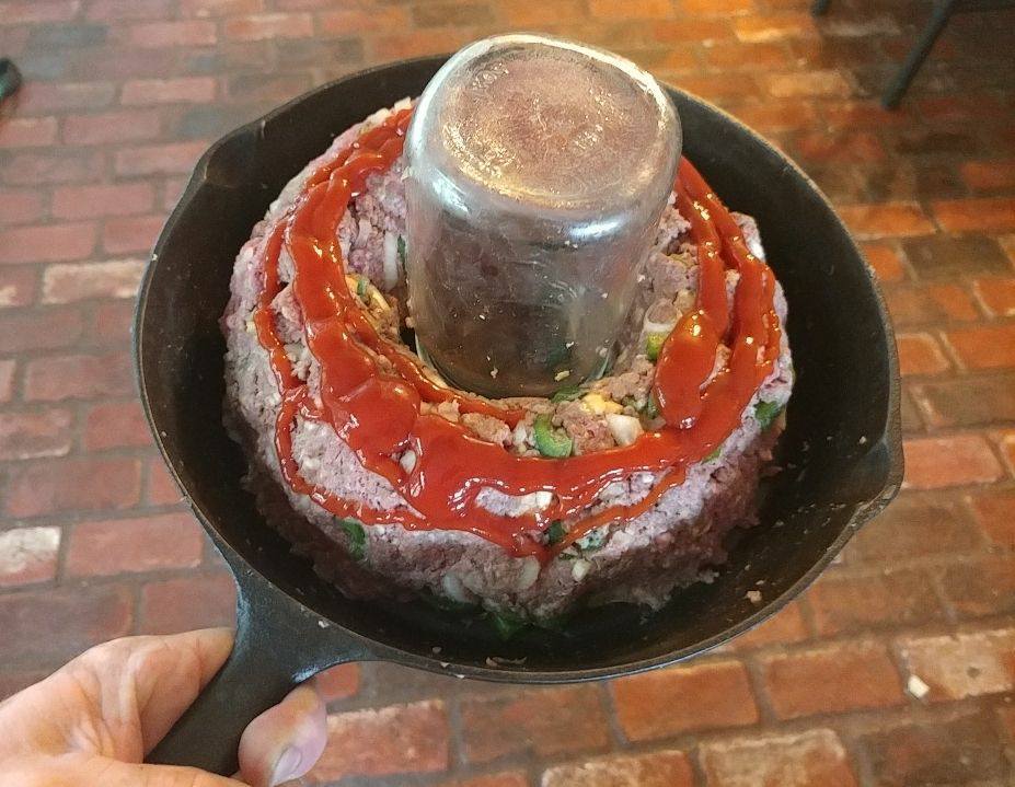 Raw meatloaf shaped around a jar in cast iron skillet, topped with Sriracha ketchup