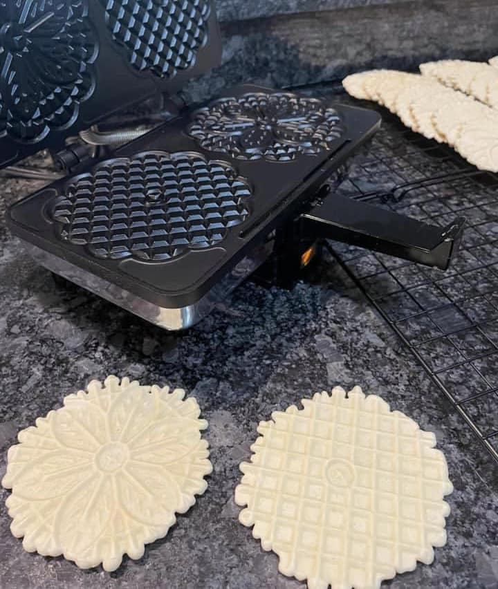 open pizzelle maker with patterned plates and freshly cooked pizzelles beside it