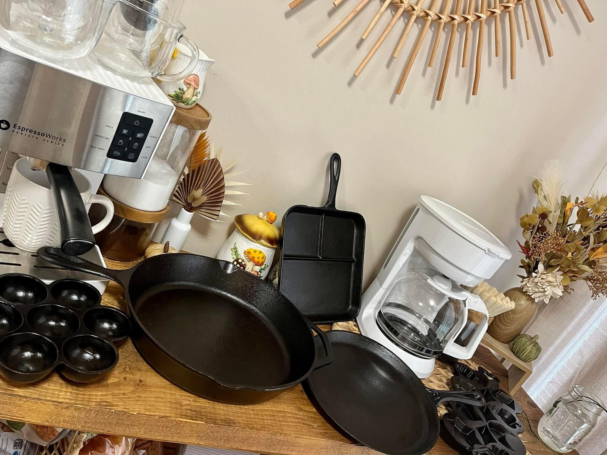 Beautifully restored vintage cast iron pans and skillets arranged on wooden counter with coffee maker and autumn decor