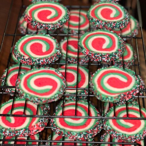 A wire rack filled with baked Christmas pinwheel sugar cookies featuring distinct red, white, and green swirls, with edges coated in holiday sprinkles.