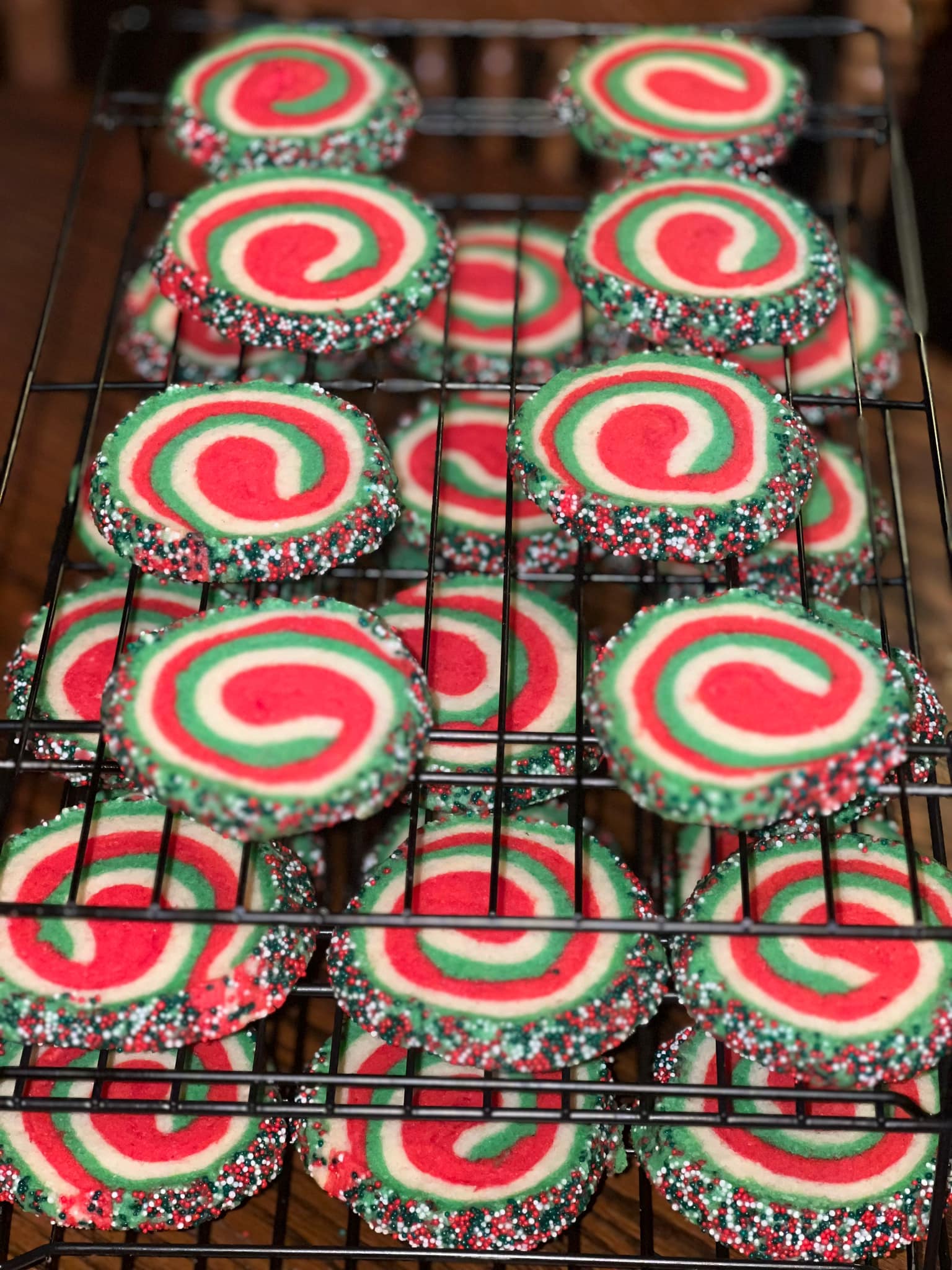 A wire rack filled with baked Christmas pinwheel sugar cookies featuring distinct red, white, and green swirls, with edges coated in holiday sprinkles.