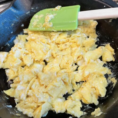 Fluffy yellow scrambled eggs being folded in a cast iron skillet with a green silicone spatula, showing distinct curds and no sticking to the pan