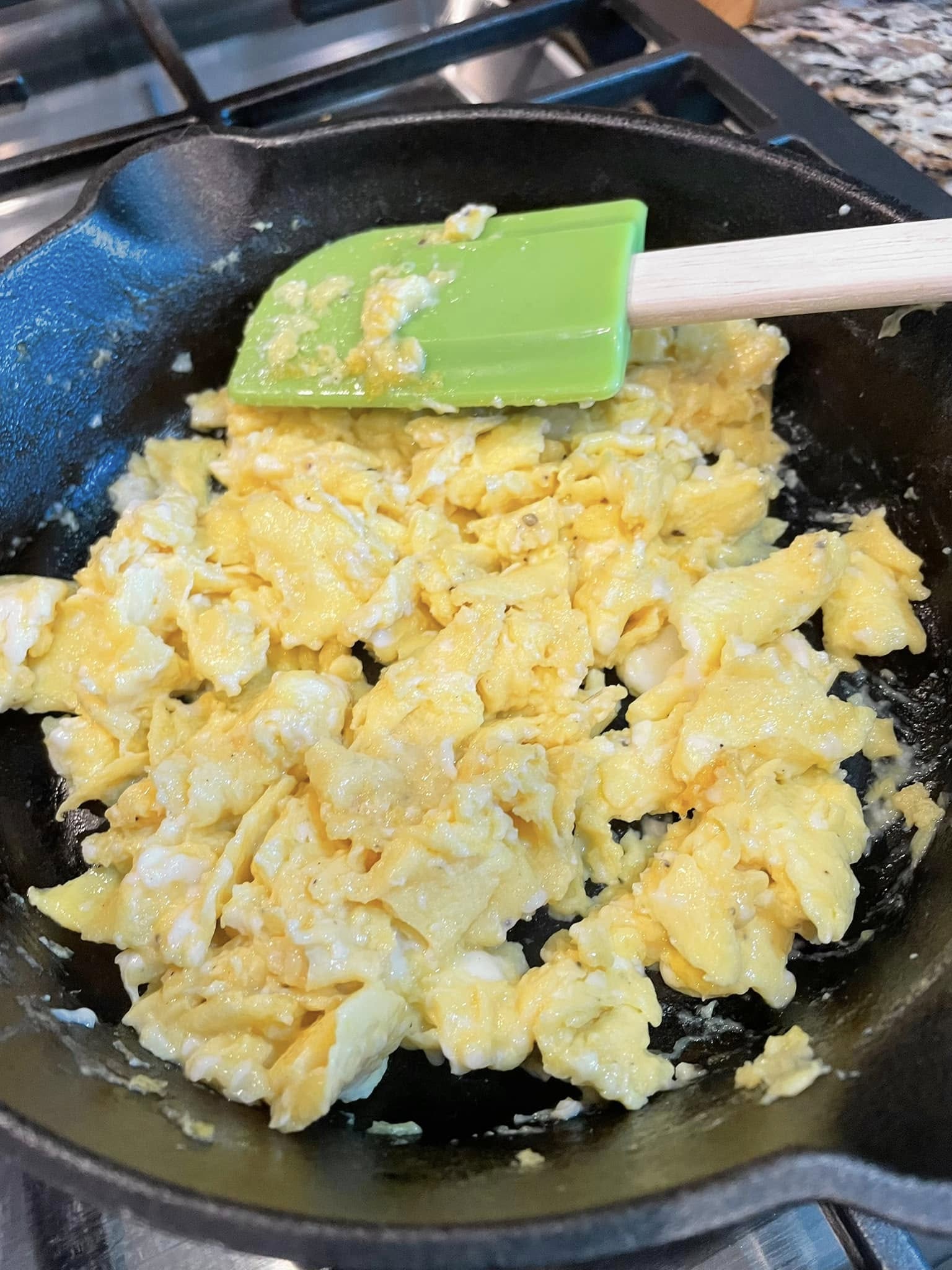 Fluffy yellow scrambled eggs being folded in a cast iron skillet with a green silicone spatula, showing distinct curds and no sticking to the pan