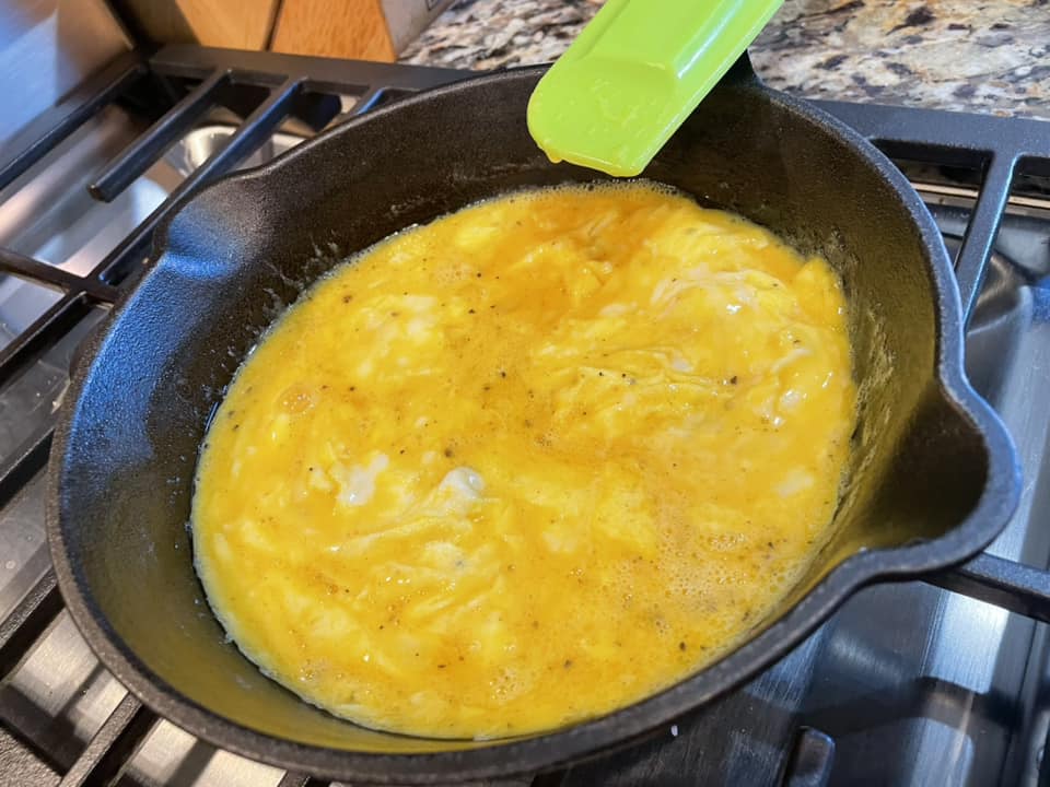 Raw scrambled eggs poured into a hot cast iron skillet with melted butter, beginning to set around the edges, with a green silicone spatula resting on the rim.