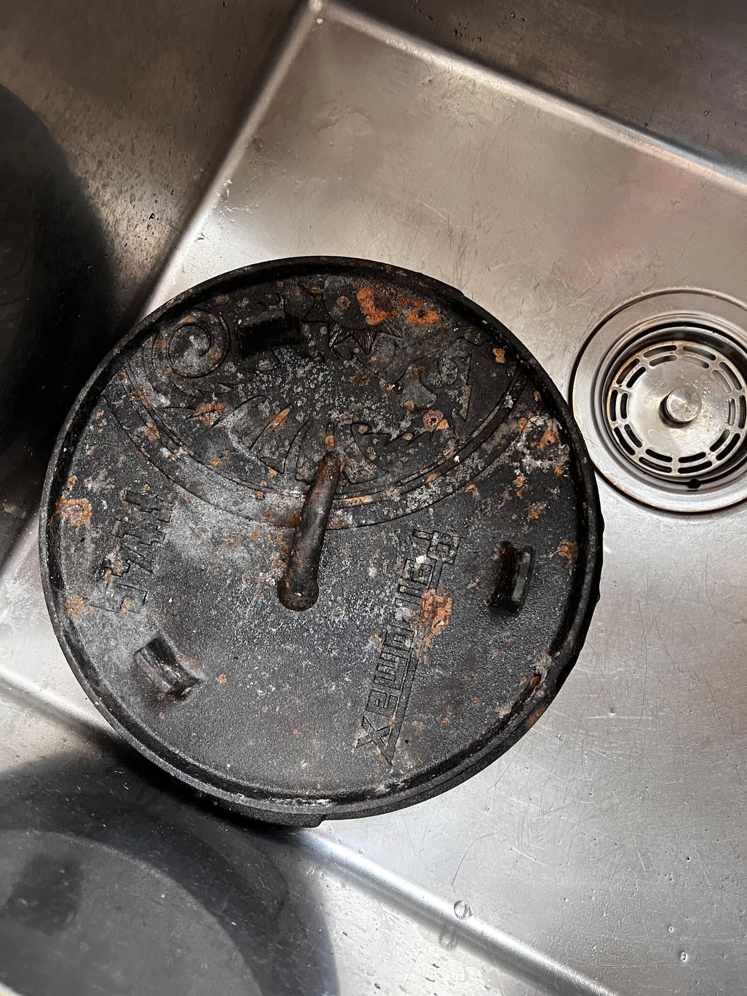 The rusted cast iron lid sitting in a stainless steel sink, covered in orange rust spots and white oxidation before cleaning