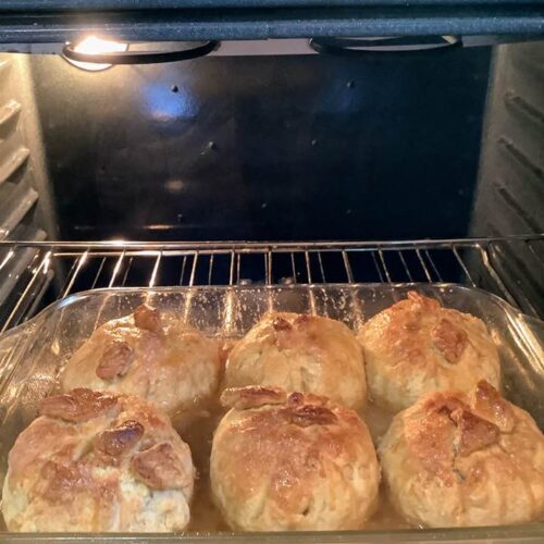 Golden brown apple dumplings baking in a clear glass dish inside an oven, surrounded by bubbling caramel sauce.