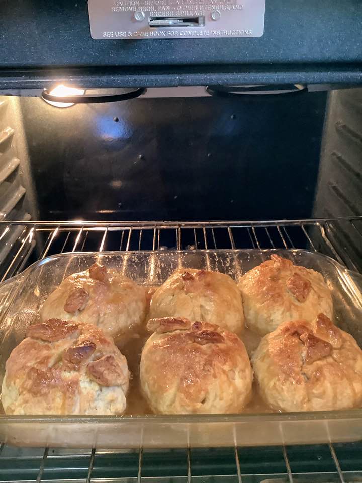 Golden brown apple dumplings baking in a clear glass dish inside an oven, surrounded by bubbling caramel sauce.