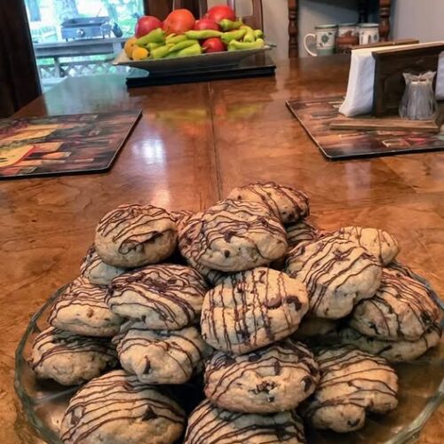 A large pile of thick, golden brown chocolate chip cookies stacked on a glass platter, decorated with a zig-zag chocolate drizzle.