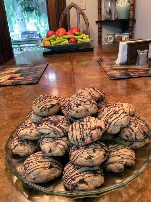 A large pile of thick, golden brown chocolate chip cookies stacked on a glass platter, decorated with a zig-zag chocolate drizzle.