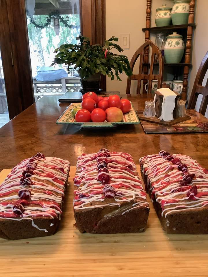 Three loaves of cranberry bread lined up on a wooden board, decorated with white icing drizzle, red icing drizzle, and topped with sugared cranberries.
